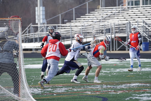 A group of young men playing a game of lacrosse on a field with some snow patches. Players are wearing helmets and jerseys of different colors, with some actively engaged in running or defending. The background shows empty bleachers and an overcast sky.