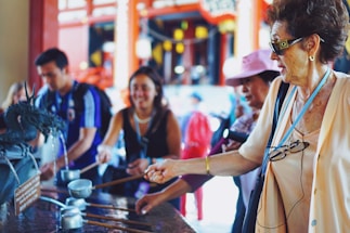 People are gathered around a traditional water fountain, performing a ritual cleansing with ladles. The group appears to be diverse in age and ethnicity, engaging in a cultural activity. The background features an ornate, vibrant architecture, suggesting a temple setting.