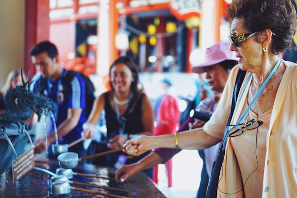 People are gathered around a traditional water fountain, performing a ritual cleansing with ladles. The group appears to be diverse in age and ethnicity, engaging in a cultural activity. The background features an ornate, vibrant architecture, suggesting a temple setting.