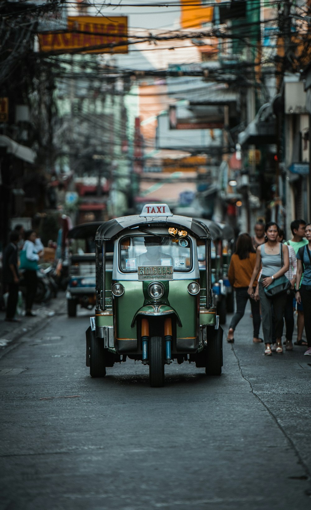 green auto rickshaw on road in between buildings with people walking around during daytime