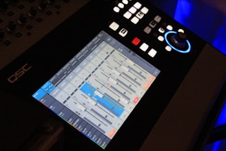 A digital audio mixing console with a touchscreen interface displaying various sliders and controls for audio management. The touchscreen shows multiple channels with sliders, buttons, and meters, some highlighted in blue. Above the screen, there are several physical buttons and a rotary knob with a blue light around it.