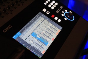 A digital audio mixing console with a touchscreen interface displaying various sliders and controls for audio management. The touchscreen shows multiple channels with sliders, buttons, and meters, some highlighted in blue. Above the screen, there are several physical buttons and a rotary knob with a blue light around it.