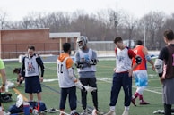 A group of young men on a sports field wearing lacrosse gear, including jerseys and pads, appear to be in a casual meeting or warm-up session. Some players are holding lacrosse sticks, and their attire suggests they are part of a sports team. The setting is outdoors, possibly in a school or community field, with a brick building and bare trees in the background.