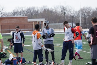 A group of young men on a sports field wearing lacrosse gear, including jerseys and pads, appear to be in a casual meeting or warm-up session. Some players are holding lacrosse sticks, and their attire suggests they are part of a sports team. The setting is outdoors, possibly in a school or community field, with a brick building and bare trees in the background.