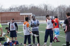 A group of young men on a sports field wearing lacrosse gear, including jerseys and pads, appear to be in a casual meeting or warm-up session. Some players are holding lacrosse sticks, and their attire suggests they are part of a sports team. The setting is outdoors, possibly in a school or community field, with a brick building and bare trees in the background.