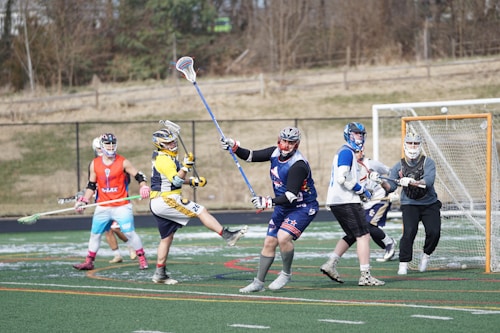 A group of lacrosse players engaged in a game on a turf field. The players are wearing protective gear including helmets and gloves, and are holding lacrosse sticks. The scene captures dynamic movement, with one player in mid-action attempting to score a goal, while a goalie stands in front of the net. Other players are observing or preparing for their next move. The field is marked with various sports lines, and a fence with a wooded area is visible in the background.