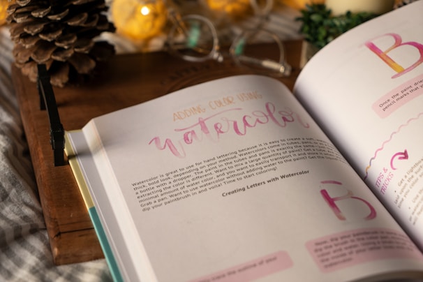 An open book displaying pages about watercolor techniques. The page headings and some text are in varied colors, evoking a creative theme. The background features a rustic wooden surface, with a pine cone, warm-toned decorative lights, and a partially visible green plant.