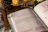 An open book displaying pages about watercolor techniques. The page headings and some text are in varied colors, evoking a creative theme. The background features a rustic wooden surface, with a pine cone, warm-toned decorative lights, and a partially visible green plant.