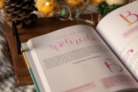An open book displaying pages about watercolor techniques. The page headings and some text are in varied colors, evoking a creative theme. The background features a rustic wooden surface, with a pine cone, warm-toned decorative lights, and a partially visible green plant.