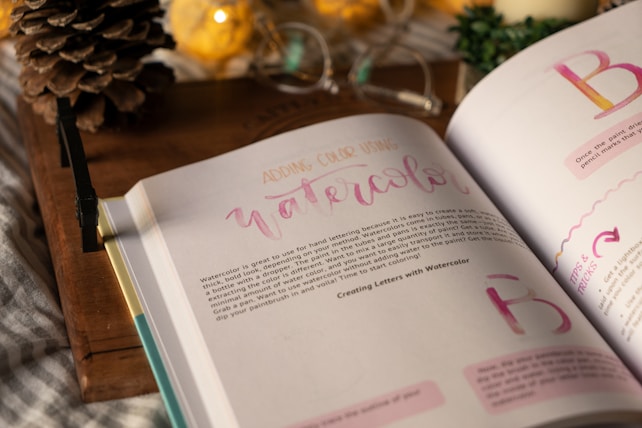 An open book displaying pages about watercolor techniques. The page headings and some text are in varied colors, evoking a creative theme. The background features a rustic wooden surface, with a pine cone, warm-toned decorative lights, and a partially visible green plant.