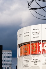 A large, cylindrical structure displays time zones for various cities around the world, with numbers and city names on its surface. The sky is overcast, providing a dramatic backdrop, while the structure itself features a globe-like design at the top.