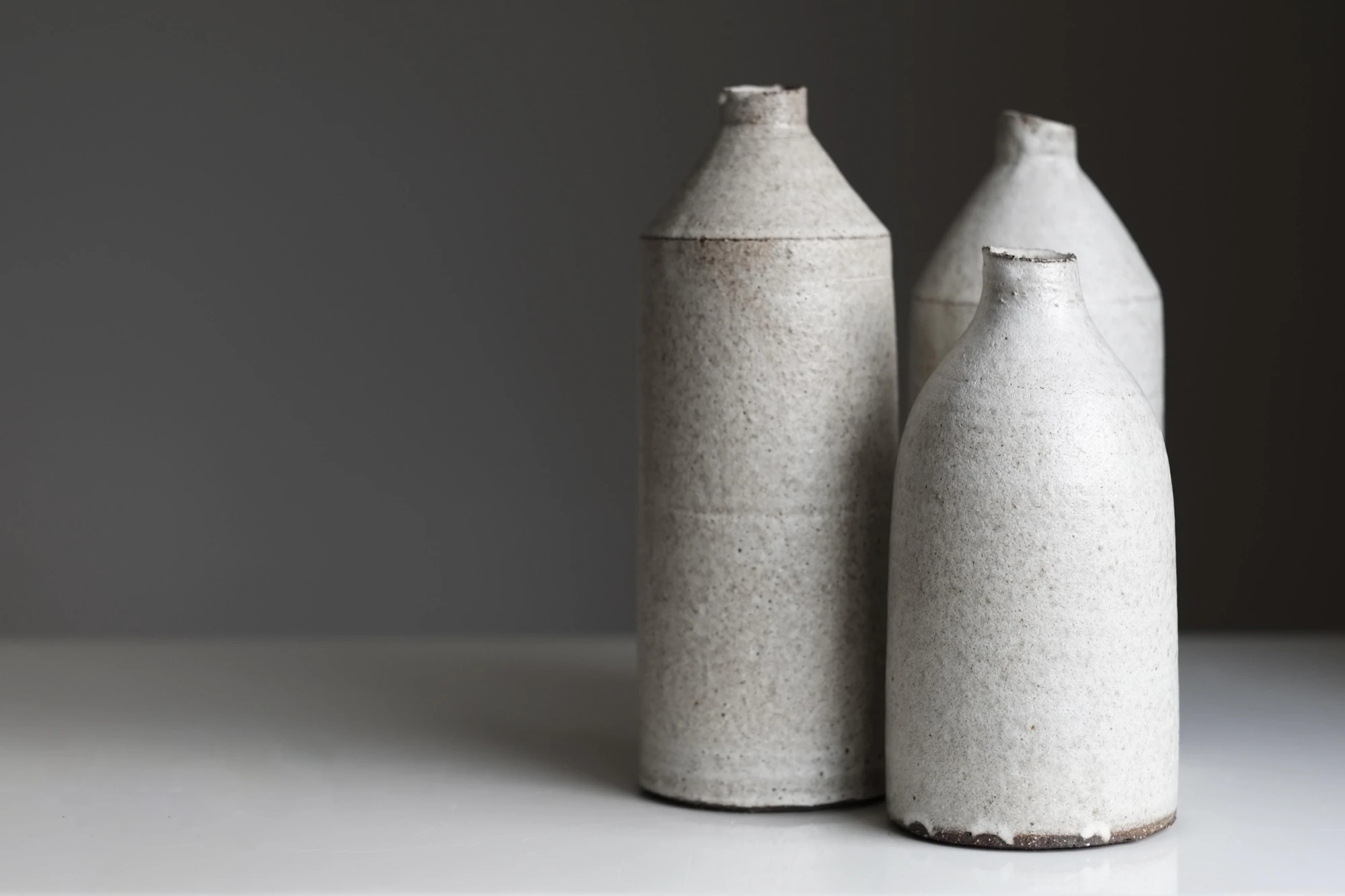 Three tall speckled stoneware bottles standing on a white table against a charcoal wall