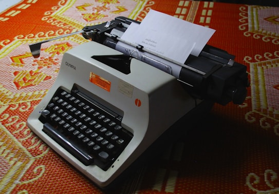 A vintage typewriter with a sheet of paper inserted, placed on a colorful, patterned fabric. The typewriter has a beige body with black keys, and visible brand name 'Olympia'. The fabric underneath features intricate designs in orange, white, and pink colors.