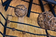 A series of spherical wicker lampshades hang from the ceiling, which is made of straw with wooden beams intersecting across it. The intricate weaving of the lampshades contrasts with the natural texture of the straw, creating an earthy and rustic aesthetic.