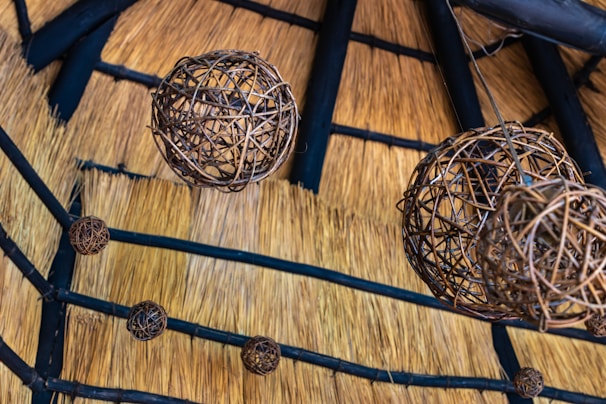 A series of spherical wicker lampshades hang from the ceiling, which is made of straw with wooden beams intersecting across it. The intricate weaving of the lampshades contrasts with the natural texture of the straw, creating an earthy and rustic aesthetic.