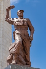 A bronze statue of a soldier or revolutionary figure is depicted in a dynamic pose. The figure is wearing a cap and military-style clothing, holding an object raised in one hand. A tall, rectangular stone structure is visible in the background, contrasting against the clear blue sky.