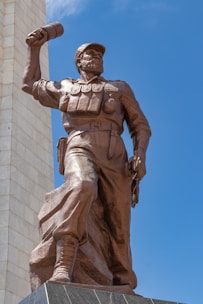 A bronze statue of a soldier or revolutionary figure is depicted in a dynamic pose. The figure is wearing a cap and military-style clothing, holding an object raised in one hand. A tall, rectangular stone structure is visible in the background, contrasting against the clear blue sky.