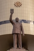 A bronze statue of a man in a suit holding a book aloft, placed in front of a tiled building. Above the statue is a sun emblem with a stylized bull's head in the center.