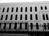 A black and white photograph of a building with architectural features resembling traditional Islamic design. The facade includes multiple rows of tall, narrow windows with an intricate metal fence in front of it. The building displays decorative elements along the edges of the windows and a scalloped pattern along the roofline.