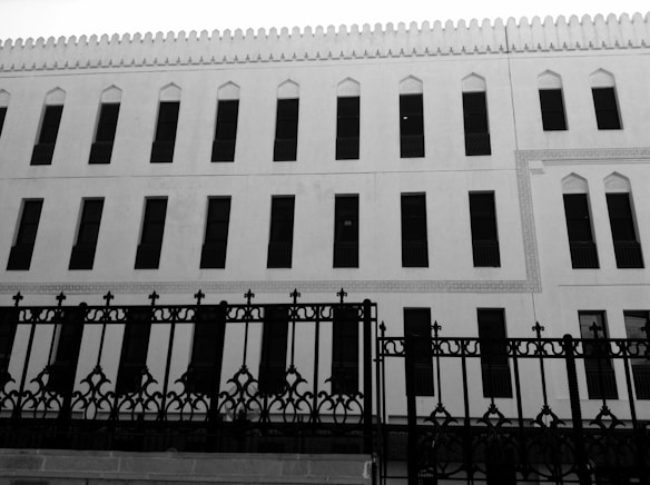A black and white photograph of a building with architectural features resembling traditional Islamic design. The facade includes multiple rows of tall, narrow windows with an intricate metal fence in front of it. The building displays decorative elements along the edges of the windows and a scalloped pattern along the roofline.