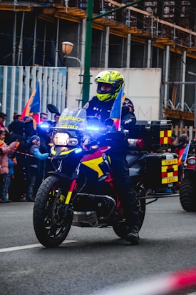 A motorcyclist is part of a parade or public event, wearing a bright neon yellow helmet and dark protective gear. The motorcycle is equipped with emergency lights, which are shining blue. It displays flags on the front and has markings that suggest it belongs to an emergency services department.