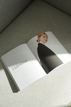 An open magazine lies on a light-colored textured surface. The left page features the word 'Valentine' in elegant font, with some smaller text beneath it. The right page displays a portrait of a person with red hair pulled back, wearing a black jacket. Soft lighting creates gentle shadows.