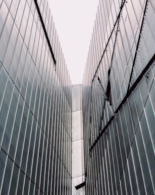 Tall metallic walls converge towards the sky, forming a narrow passageway. The surface is covered in evenly spaced vertical panels that reflect the light, creating a modern and industrial appearance.