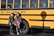 Two people are standing in front of a yellow school bus with the text 'County Career & Technical Center' written on its side. Both individuals are wearing casual clothing and are posed next to the bus shadow on the ground.