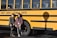 Two people are standing in front of a yellow school bus with the text 'County Career & Technical Center' written on its side. Both individuals are wearing casual clothing and are posed next to the bus shadow on the ground.