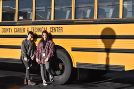Two people are standing in front of a yellow school bus with the text 'County Career & Technical Center' written on its side. Both individuals are wearing casual clothing and are posed next to the bus shadow on the ground.
