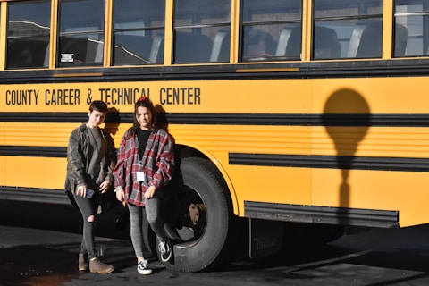 Two people are standing in front of a yellow school bus with the text 'County Career & Technical Center' written on its side. Both individuals are wearing casual clothing and are posed next to the bus shadow on the ground.