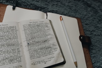 An open book with text written in a foreign script lies on a notebook, next to a pencil. The notebook is bound in a brown leather cover, and the setting appears to be a soft, textured surface.