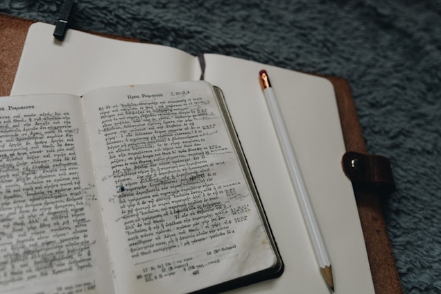 An open book with text written in a foreign script lies on a notebook, next to a pencil. The notebook is bound in a brown leather cover, and the setting appears to be a soft, textured surface.