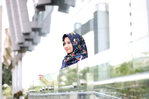 A woman wearing a floral-patterned hijab stands by a glass railing in a modern, urban environment. The background includes architectural structures with reflections and blurred greenery.