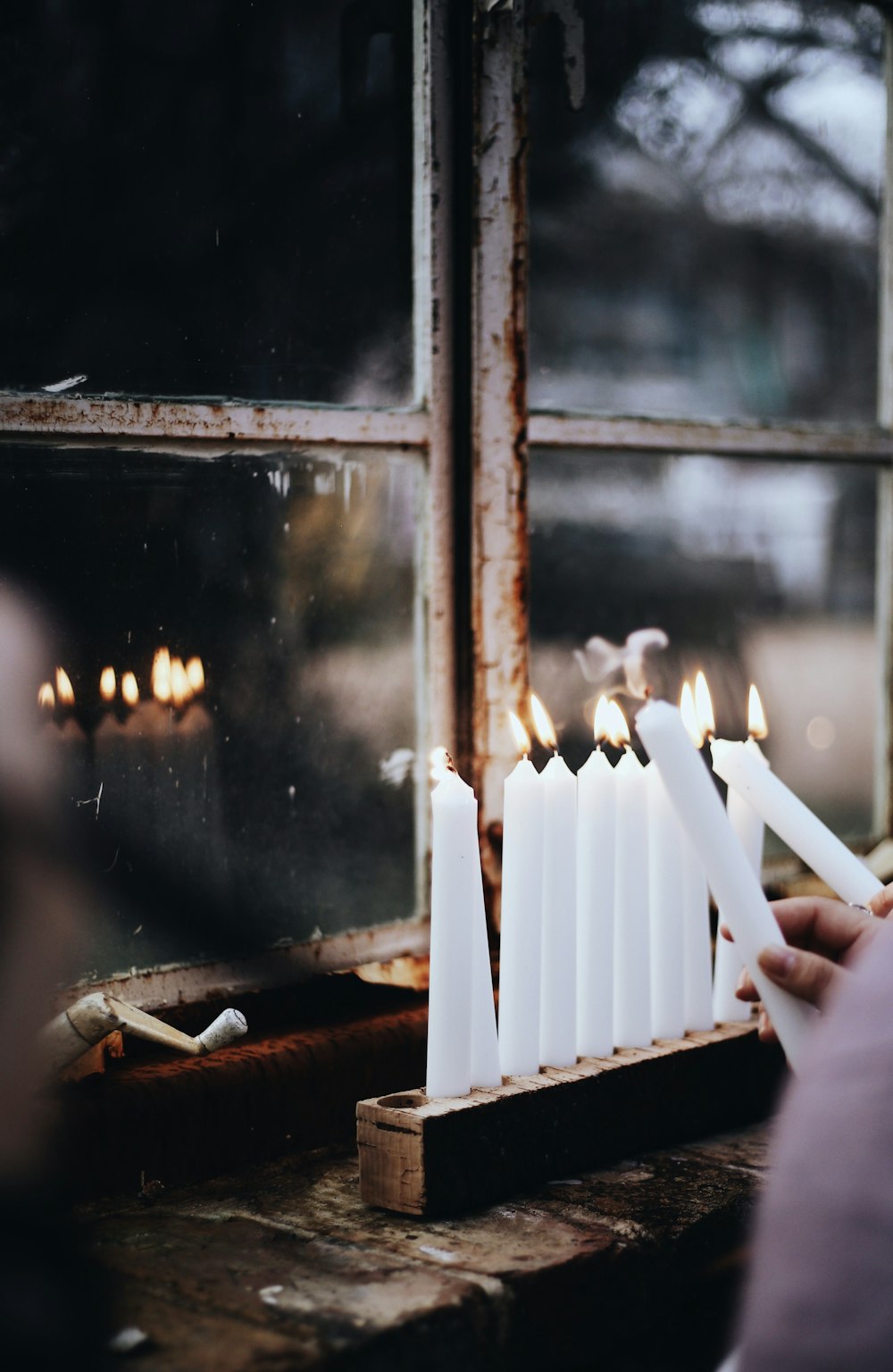 person lighting white candlesticks in a rack near glass windowpane