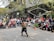 A street performer is entertaining a crowd with a dynamic display using a large, rectangular frame. The performer, who is shirtless and wearing black pants, has captured the attention of a sizable audience of various ages. The scene takes place on a city street surrounded by modern buildings and trees with autumn leaves. Many spectators are standing or sitting on the sidewalk, watching intently.