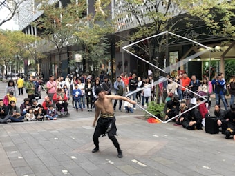 A street performer is entertaining a crowd with a dynamic display using a large, rectangular frame. The performer, who is shirtless and wearing black pants, has captured the attention of a sizable audience of various ages. The scene takes place on a city street surrounded by modern buildings and trees with autumn leaves. Many spectators are standing or sitting on the sidewalk, watching intently.