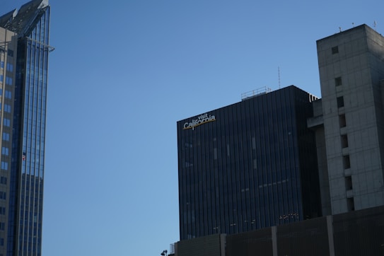 A skyline with modern office buildings under a clear blue sky. One building has reflective glass panels, and another features a sign that reads 'visit California'. The structures are primarily vertical with different textures and designs.