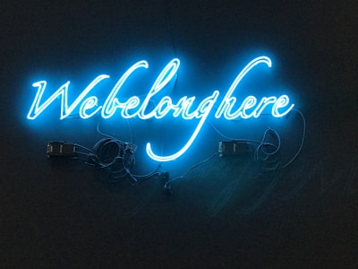 A neon sign with the phrase 'We belong here' written in bright blue cursive script against a dark background. The sign is illuminated, casting a soft glow, with visible wiring connected to the letters.