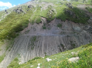 A steep, rugged hillside with sparse vegetation and visible erosion patterns. The slope is covered in patches of green grass and scattered rocks. There is a contrast between the grassy areas and the bare, eroded sections with shallow grooves.