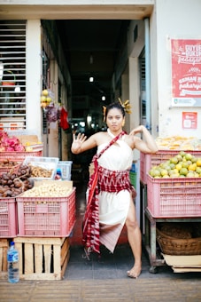 A woman dressed in traditional clothing poses in front of a market stall. The stall is filled with a variety of fruits and vegetables in crates, such as mangoes and potatoes. The market has a vibrant and bustling atmosphere, with signs in the background and items hanging above.