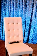 A white upholstered chair with a quilted design is set against a textured blue curtain background. A microphone rests on the seat, suggesting a setting prepared for a presentation or speech.