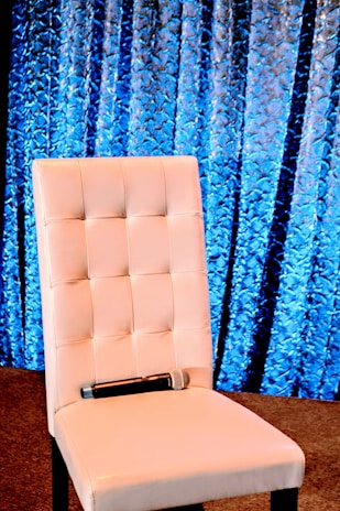 A white upholstered chair with a quilted design is set against a textured blue curtain background. A microphone rests on the seat, suggesting a setting prepared for a presentation or speech.