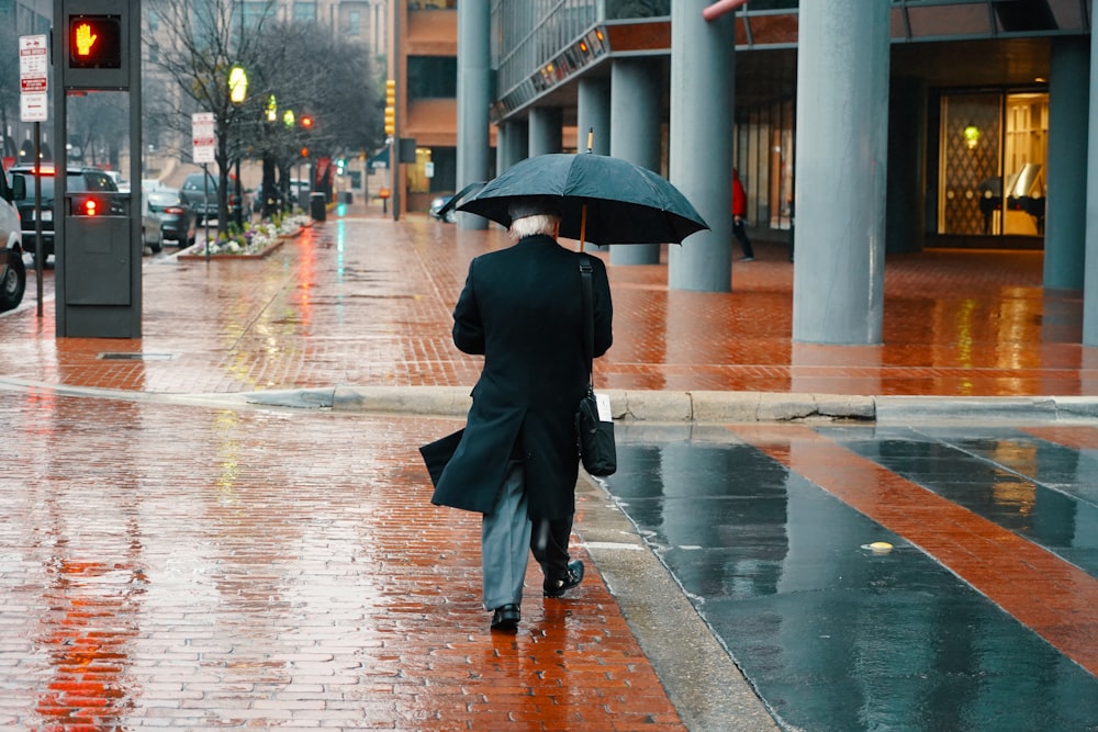 man wearing black coat, gray dress pants holding umbrella walking on wet street