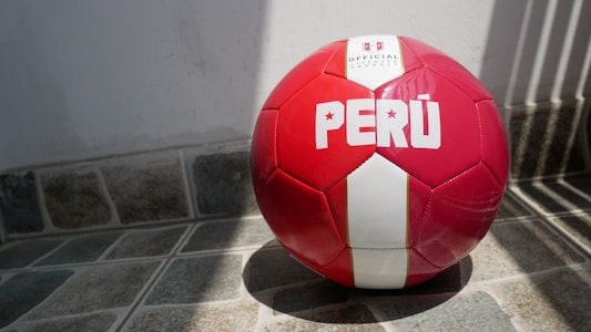A red and white soccer ball with the word 'PERÚ' printed on it rests on a tiled floor with shadows cast from nearby bars.