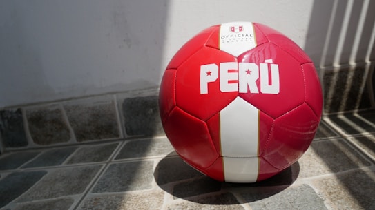 A red and white soccer ball with the word 'PER&Uacute;' printed on it rests on a tiled floor with shadows cast from nearby bars.