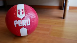 A red and white soccer ball featuring the word 'PERÚ' and the emblem with 'FPF' is placed on a wooden floor inside a room. The ball's glossy surface reflects some light, and there is a piece of furniture in the background.