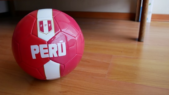 A red and white soccer ball featuring the word 'PER&Uacute;' and the emblem with 'FPF' is placed on a wooden floor inside a room. The ball's glossy surface reflects some light, and there is a piece of furniture in the background.