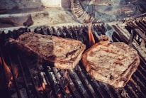 Two juicy steaks sizzle on a charcoal grill, with flames and smoke rising around them. The steaks have a charred, seared surface, indicative of being cooked to perfection.