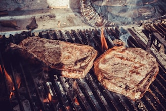 Two juicy steaks sizzle on a charcoal grill, with flames and smoke rising around them. The steaks have a charred, seared surface, indicative of being cooked to perfection.